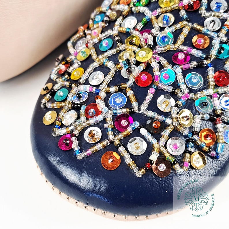 May include: Close-up of a navy blue leather slipper with colorful sequins and beads. The slipper has a geometric pattern and is decorated with a variety of colors, including gold, silver, red, green, blue, and purple.