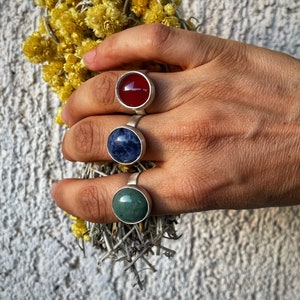 May include: Three silver rings with different colored stones. The ring on the index finger has a red stone, the middle finger has a blue stone, and the ring finger has a green stone.