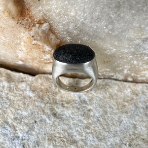 May include: A silver ring with a dark, textured stone set in a brushed metal band. The stone is oval-shaped and appears to be a dark gray or black color. The ring is displayed on a light-colored, textured surface.