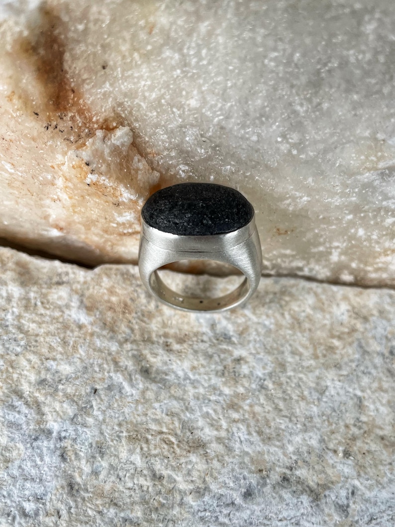 May include: A silver ring with a dark, textured stone set in a brushed metal band. The stone is oval-shaped and appears to be a dark gray or black color. The ring is displayed on a light-colored, textured surface.