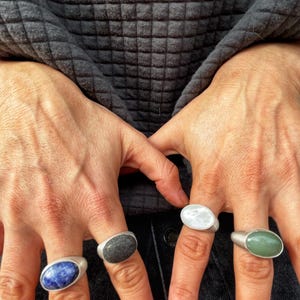 May include: Four silver rings, each with an oval-shaped stone in different colors: blue, gray, white, and green. The rings are displayed on fingers against a dark gray textured background.
