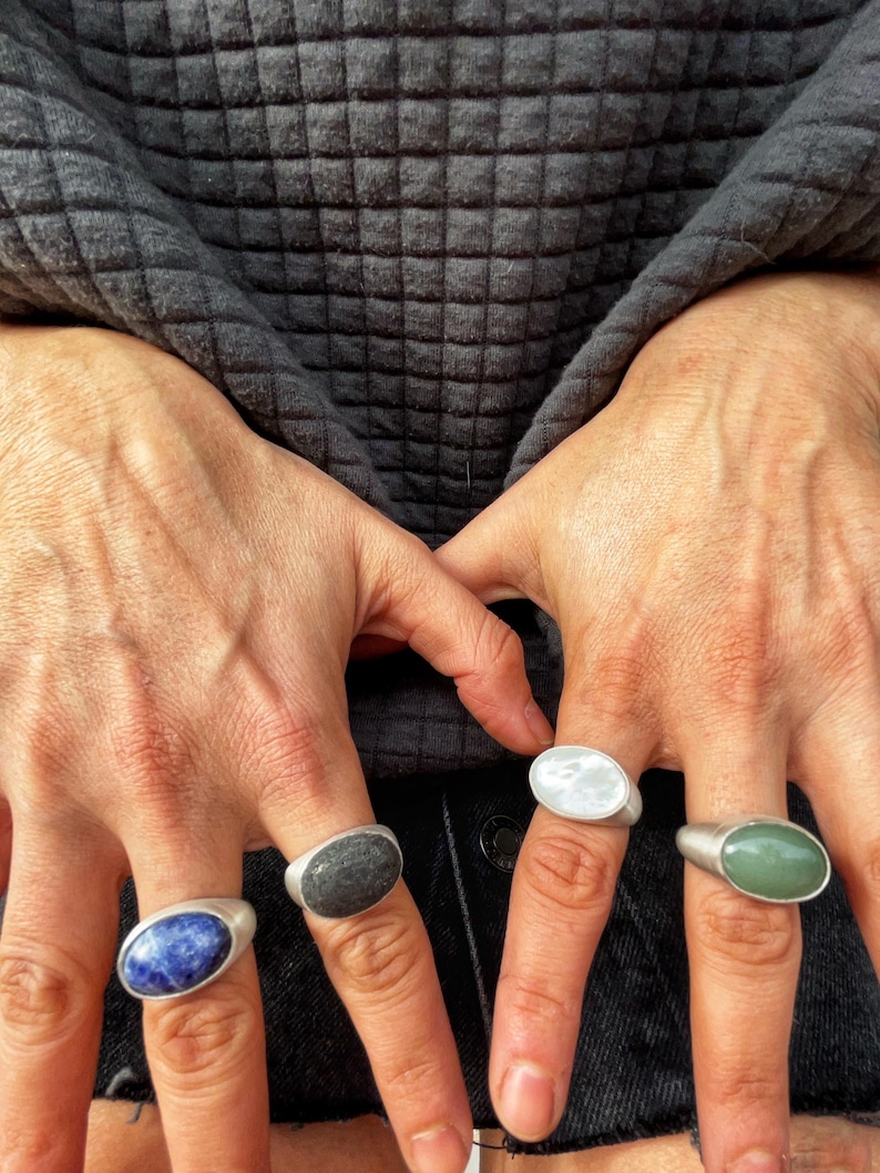 May include: Four silver rings, each with an oval-shaped stone in different colors: blue, gray, white, and green. The rings are displayed on fingers against a dark gray textured background.