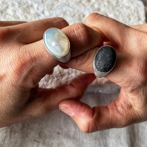 May include: Two rings with oval-shaped stones. One ring features a pearlescent white stone, and the other has a black, textured stone. Both rings have a silver band.