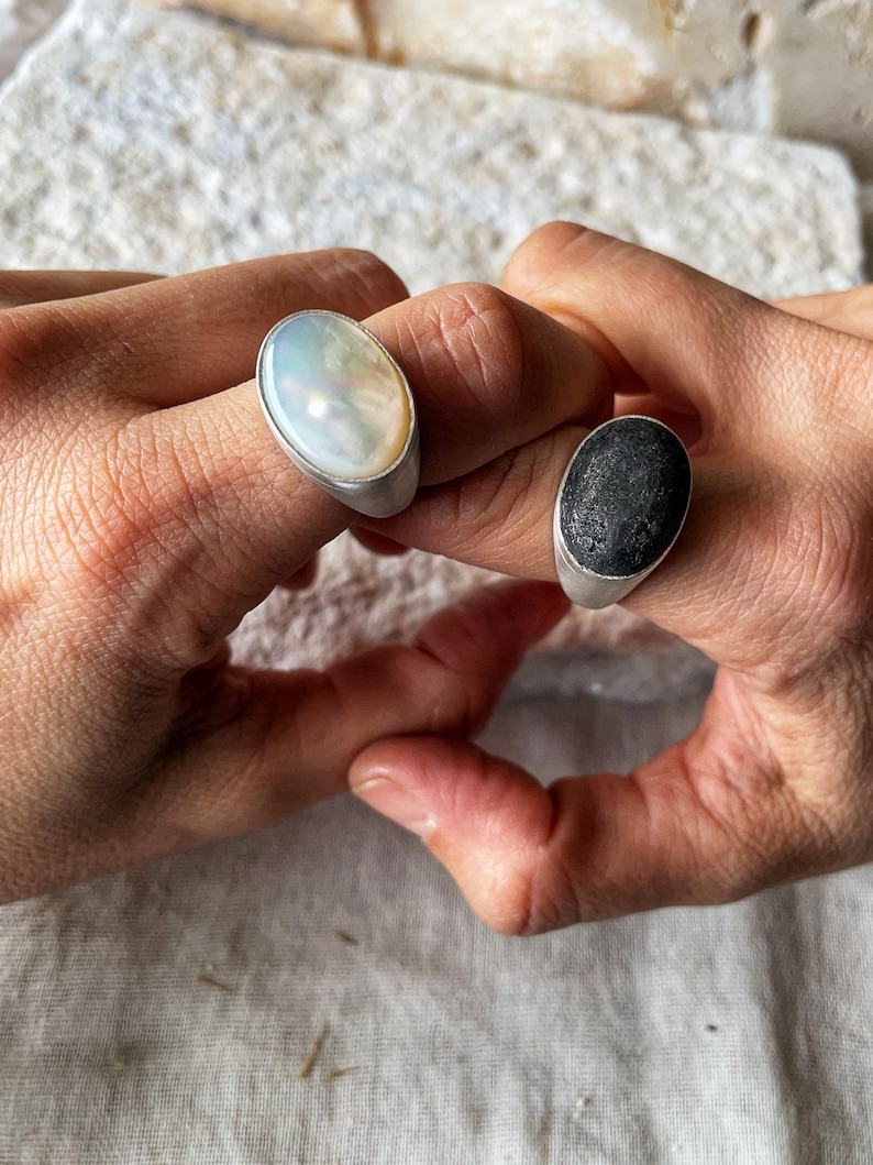 May include: Two rings with oval-shaped stones. One ring features a pearlescent white stone, and the other has a black, textured stone. Both rings have a silver band.