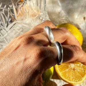 May include: Two silver rings worn on a finger. The rings are smooth and have a rounded shape. The rings are worn on a hand with a light brown skin tone.