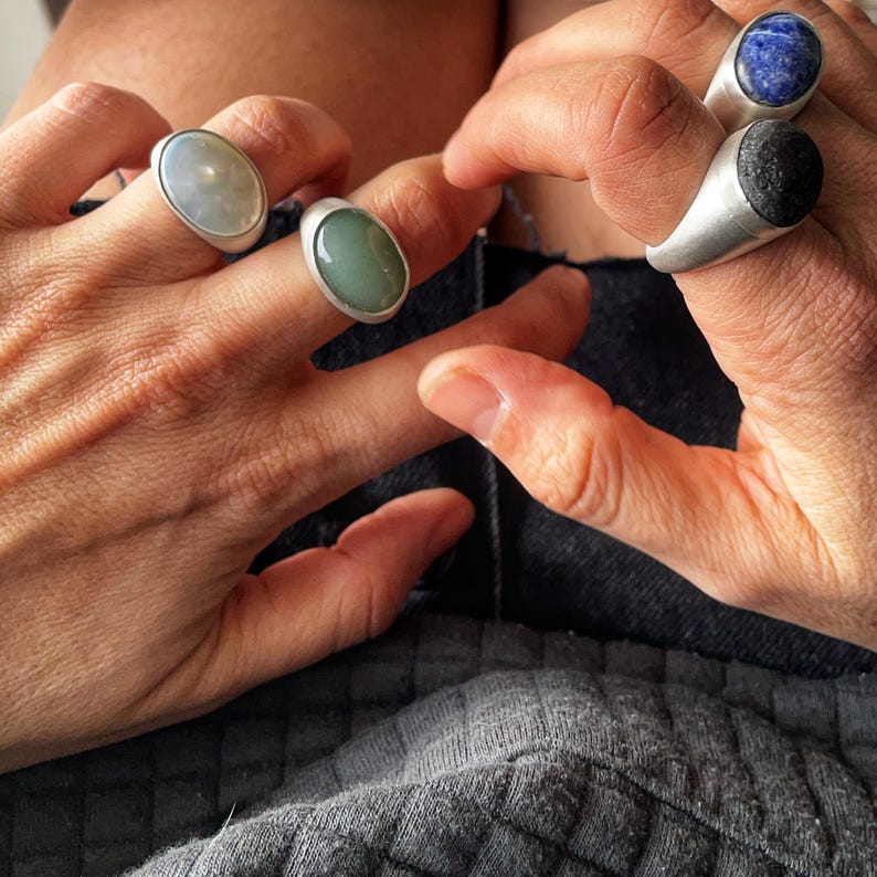 May include: Four silver rings with oval-shaped gemstones in various colors: light green, dark green, blue, and black. The rings are displayed on fingers, showcasing the jewelry's design and colors. The gemstones are set in a simple, modern style.