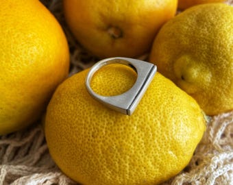 May include: A silver ring with a unique, geometric design sits on top of a yellow lemon. The lemon is surrounded by other lemons and a white crocheted surface.