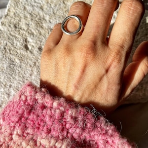 May include: A silver ring with a large, open circle design. The ring is worn on a hand with a pink and white knitted sweater.