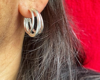 May include: Silver hoop earrings with a triple-band design. The earrings are worn on an ear, with the skin visible. The background is a solid red color. The earrings are a simple, elegant accessory.