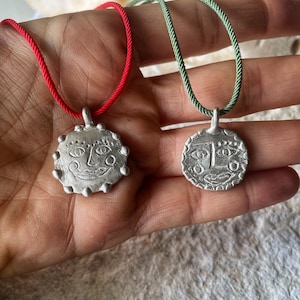 以下が含まれることがあります： 顔の形をしたペンダントが付いた銀のネックレスが2本。1本のネックレスは赤い紐、もう1本のネックレスは緑の紐です。