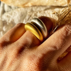 以下が含まれることがあります： 指に重ねられた2つのシンプルでモダンなリング。上のリングはシルバー、下のリングはゴールドです。