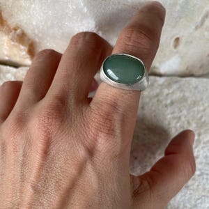 May include: A silver ring with a large, oval, light green stone. The ring has a simple, modern design with a brushed metal finish. The stone is set in a bezel setting, and the ring is shown on a finger.