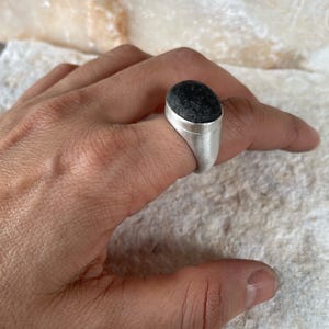 May include: A silver ring with a dark, textured, oval-shaped stone. The ring is set in a simple, modern style, showcasing the stone's natural appearance. The ring is shown on a hand.
