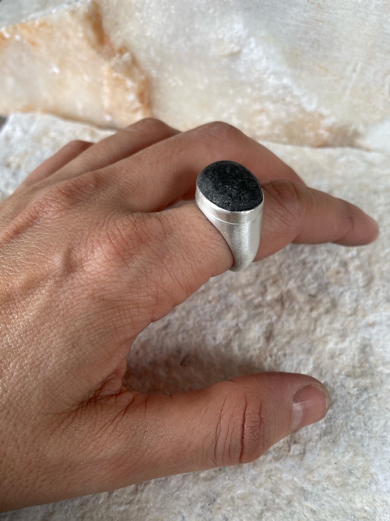 May include: A silver ring with a dark, textured, oval-shaped stone. The ring is set in a simple, modern style, showcasing the stone's natural appearance. The ring is shown on a hand.