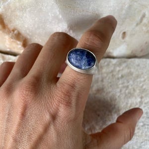 May include: A silver ring featuring a large, oval-shaped, deep blue gemstone with white marbling. The ring has a simple, modern design with a wide band. The gemstone is set in a bezel setting, and the ring is shown on a finger.
