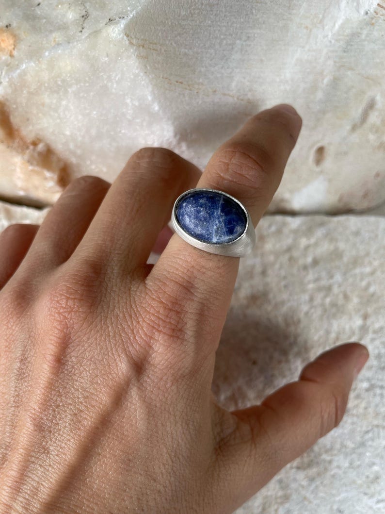 May include: A silver ring featuring a large, oval-shaped, deep blue gemstone with white marbling. The ring has a simple, modern design with a wide band. The gemstone is set in a bezel setting, and the ring is shown on a finger.