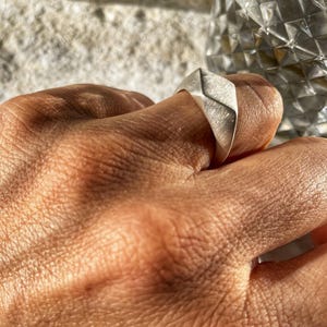 Könnte beinhalten: Ein silberner Ring mit geometrischem Design. Der Ring wird an einem Finger getragen.