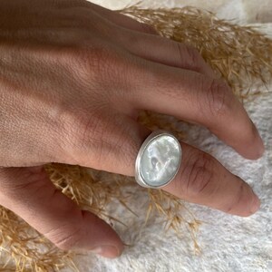 May include: A silver ring with an oval, iridescent, mother-of-pearl stone. The ring is on a finger, with a neutral background. The ring's setting is a simple, polished silver band.