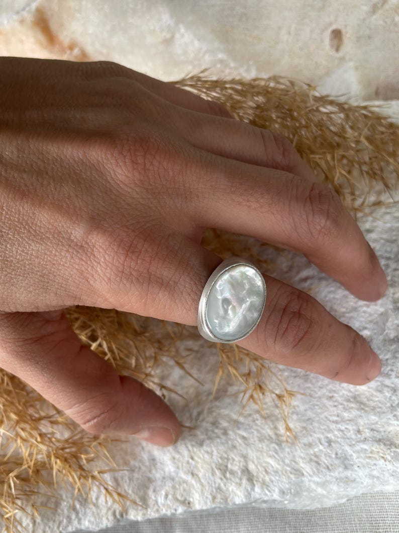 May include: A silver ring with an oval, iridescent, mother-of-pearl stone. The ring is on a finger, with a neutral background. The ring's setting is a simple, polished silver band.