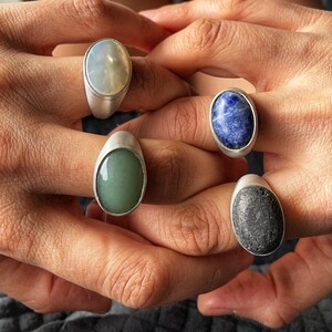 May include: Five silver rings with oval-shaped gemstones in various colors: pale green, blue, gray, and a light, iridescent color. The rings are displayed on fingers, showcasing the jewelry's design and colors.
