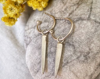 May include: A pair of silver hoop earrings with a pointed silver pendant hanging from each hoop. The earrings are resting on a gray stone.