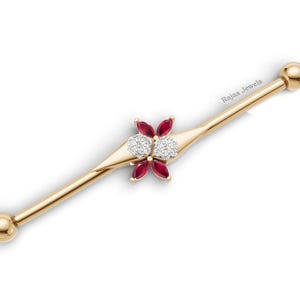 May include: A gold industrial barbell piercing with two round end balls. The center features a decorative element with four marquise-cut red rubies arranged in a flower pattern and two round white diamonds set in the middle. The text "Rajas Jewels" is visible.