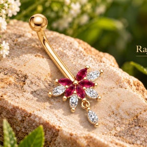 Peut inclure: Un piercing de nombril doré avec un pendentif en forme de fleur orné de pierres rouges et transparentes taille marquise, incluant une pierre pendante. Le texte "Rj Rajas Jewels" est visible dans le coin supérieur droit.