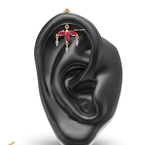 May include: A gold cartilage earring featuring three marquise-cut red gemstones arranged in a fan shape, with three small dangling clear stones. The piece includes the text Rajoo Jewels and is displayed on a black ear model.