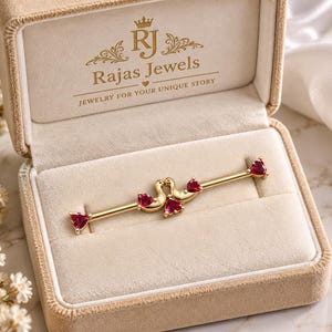 May include: A gold industrial barbell piercing with two swan figures at the center, each accented with a red heart-shaped gemstone. The barbell ends are also tipped with red heart-shaped gemstones. The jewelry is presented in a beige box.