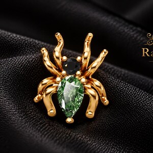 May include: A gold spider brooch featuring a pear-shaped green gemstone for the body and a round black gemstone for the head. The piece includes the RajasJewels logo with the text RJ and RajasJewels in the upper right corner of the frame.