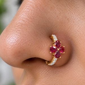 Könnte beinhalten: Ein goldener Nasenring in Creolenform mit einem vierblättrigen Blumendesign, das aus vier tropfenförmigen, tiefroten Rubinen besteht. Der Ring ist entlang der Schiene mit kleinen, klaren, rund geschliffenen weißen Steinen besetzt.