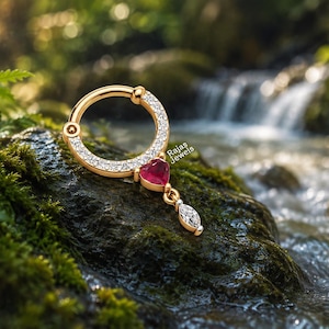 May include: A gold hoop ring featuring a row of small clear stones, a central red heart-shaped stone, and a dangling marquise-cut clear stone. The jewelry piece is marked with the text Rajas Jewels and displays a hinged closure mechanism.