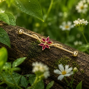 May include: A gold industrial barbell earring featuring a red flower design. The jewellery is set against a natural background of a tree branch, green leaves, and white flowers.