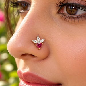 May include: A nose stud featuring a central heart-shaped red gemstone set in gold, accented by two marquise-cut clear white stones arranged in a floral-inspired pattern on a metallic base, positioned on the side of a nostril.