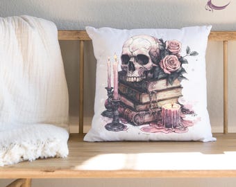 May include: A square white throw pillow featuring a printed illustration of a human skull resting on a stack of three vintage books. Beside the books are two tall pink candles in dark holders and a short pink candle with melting wax.