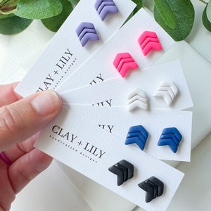 May include: Set of six pairs of handmade polymer clay earrings. Each pair features a double chevron design in a different color: purple, pink, white, blue, black, and white. The earrings are displayed on white cards with the text "CLAY + LILLY handcrafted designs".