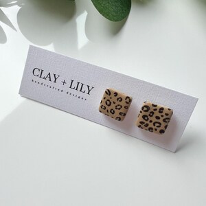 May include: Square stud earrings with a tan base and black leopard print pattern. The earrings are displayed on a white card with the text "CLAY + LILY handcrafted designs". The background is white with a green leaf.