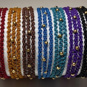 May include: A collection of seven multi-strand bracelets made of knotted cord in red, gold, brown, blue, teal, purple, and black. Each bracelet features three parallel strands woven with small, round, metallic gold beads spaced evenly throughout.