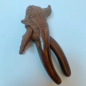 Könnte beinhalten: Ein dunkelbrauner Nussknacker aus Holz, in Form eines Vogels geschnitzt. Der Kopf, die Flügel und der Körper des Vogels sind detailliert ausgearbeitet. Die Griffe des Nussknackers sind lang und gebogen.