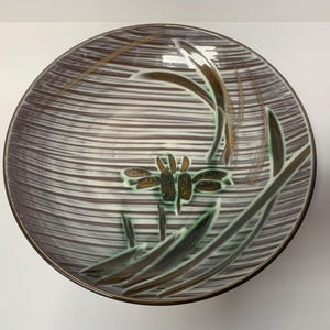 Peut inclure: Assiette décorative ronde en céramique avec un motif rayé marron et blanc. L'assiette présente un motif de libellule et de plante dans des tons marron, vert et or. Le bord est de couleur marron foncé.