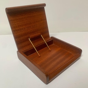 Peut inclure: Une boîte en bois avec un couvercle à charnière, ouverte pour révéler un petit compartiment. La boîte est en bois brun rougeâtre et a une finition lisse. Deux tiges métalliques dorées sont à l'intérieur de la boîte. Le couvercle est légèrement incliné.