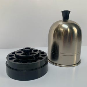May include: A stainless steel bell-shaped container with a black handle next to a black plastic multi-holed organizer. The container has a shiny, reflective surface. The organizer is round with multiple tiers of holes.