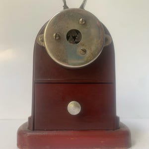 May include: A vintage pencil sharpener made of dark reddish-brown wood, with a silver-coloured metal top and two handles. It has a small drawer and a lighter red base. The sharpener is likely from the mid-20th century.