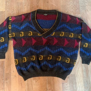 Op de afbeelding: Een vintage V-hals trui met een geometrisch patroon. De trui heeft een donkergrijze basis met blauwe, rode en gele accenten. Het ontwerp bevat zigzags, pijlen en letterachtige vormen. De kraag is gestreept met rood, blauw en geel.