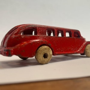 Może przedstawiać: Czerwony, metalowy autobus z białymi kołami. Autobus ma długie nadwozie i okna po bokach. Zabawka ma vintage'owy wygląd.
