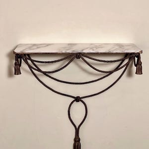 Pode incluir: Uma prateleira de parede com um tampo retangular de mármore branco com veios cinzentos. A prateleira é suportada por cordas decorativas castanho-escuras que pendem em vários festões, com borlas nas extremidades e um elemento central.