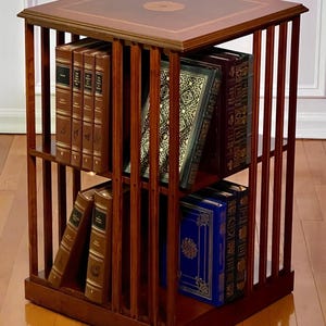 May include: A square, wooden bookcase with a dark finish, filled with books. The bookcase has vertical slats on the sides and a decorative top with a circular inlay. The books are various colors and sizes, adding to the bookcase's classic appeal.