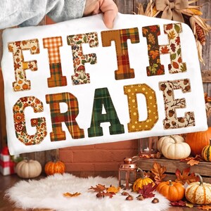 May include: A white t-shirt featuring the text FIFTH GRADE in large block letters. Each letter is filled with different autumn-themed patterns, including orange plaid, fall leaves, mushrooms, acorns, and green tartan, with visible stitching.