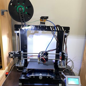 Może przedstawiać: Czarna drukarka 3D z zieloną szpulą filamentu, czarnym i srebrnym stołem roboczym oraz małym wyświetlaczem LCD z boku. Drukarka stoi na ciemnobrązowym drewnianym stole.