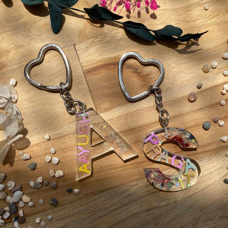 Top view of a customizable Resin Initial Keychain made with transparent epoxy resin, in which dried flowers, small stones, glitter, small leaves, etc., options can be inserted along with the name inside or without a name.